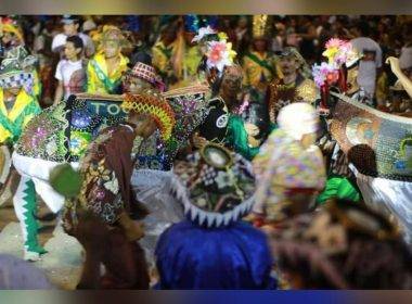 Um boneco de Bumba meu Boi está dançando em uma apresentação. Ele está todo enfeitado com brilhos, e desenhos de flores. Ao redor do boi há várias pessoas dançando com chapeis também enfeitados e coloridos.
