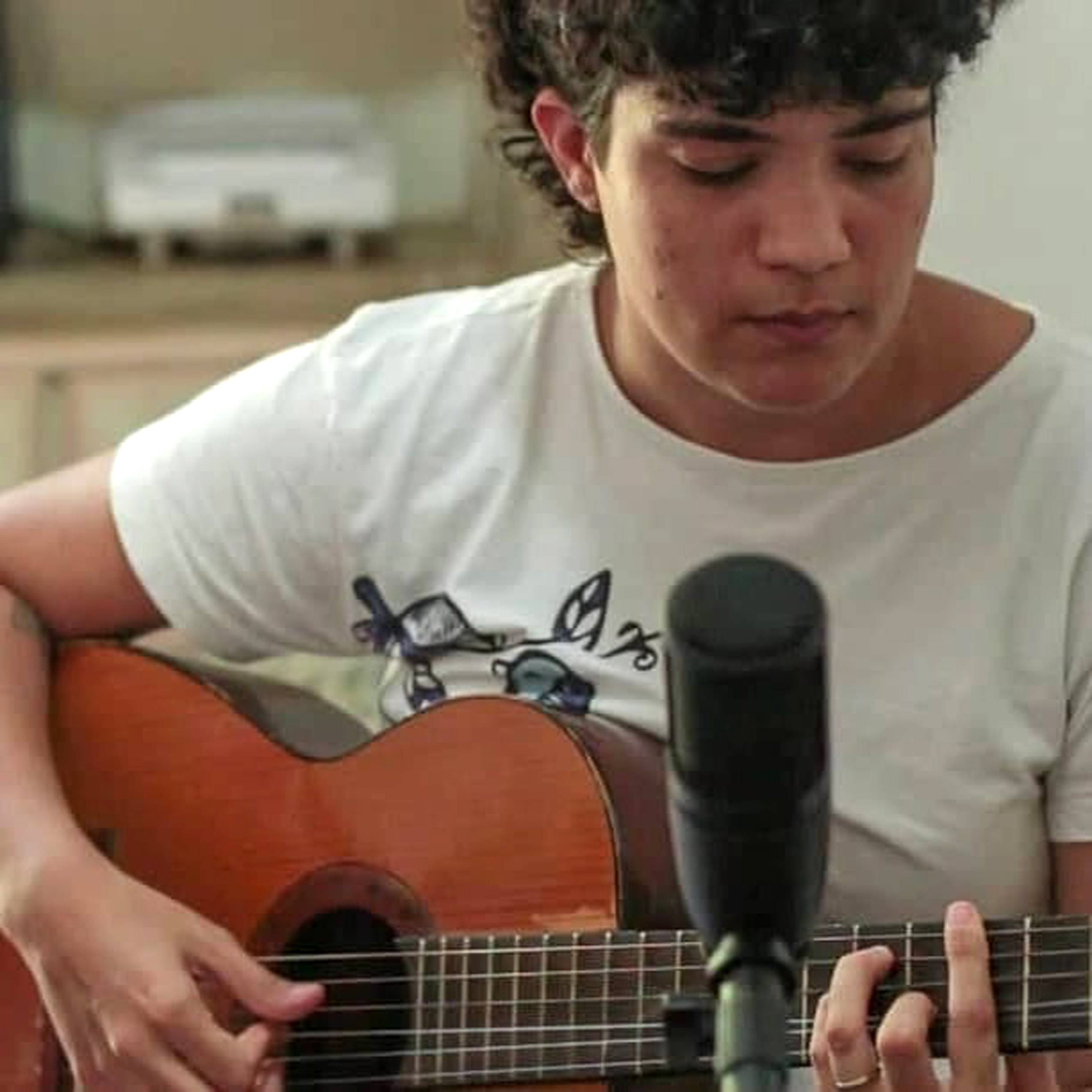 Foto de Tauana Queiroz. Com um violão nas mãos e um microfone em um tripé na frente, Tauana olha para baixo enquanto dedilha as cordas do violão. Veste uma camisa branca.