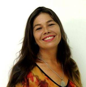 Foto de Cláudia Simone. Ela está sorrindo, usa um colar prateado com uma medalha, está vestindo uma blusa laranja e vermelha. O cabelo ondulado cai sobre os ombros.