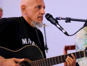 Foto de James Brito. Ele está tocando um violão e tem um microfone em um tripé na frente dele. Ele está de lado, tem uma barba branca apenas no queixo e traz a cabeça raspada. Veste uma camisa preta.