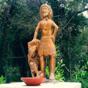 Foto de uma estátua de cor de bronze. A estátua representa um homem barbudo vestindo apenas um saiote de palha. Ele está em pé e apoia a mão em uma pequena coluna ou pilar que traz vários peixes ao seu redor.
