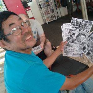 Foto de Luís Celso segurando algumas páginas finalizadas de suas histórias em quadrinhos. Ele usa óculos, veste uma camisa azul e sorri para a câmera.