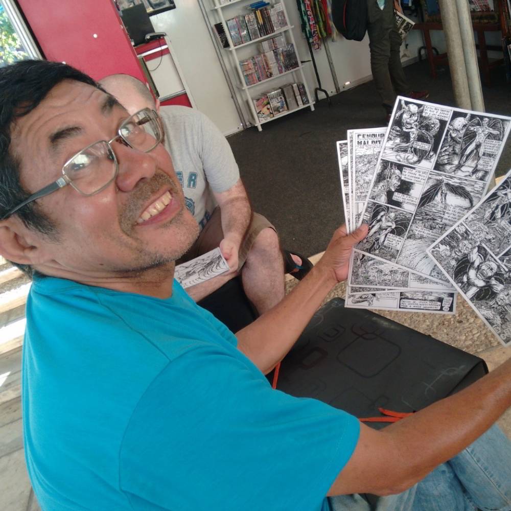Foto de Luís Celso segurando algumas páginas finalizadas de suas histórias em quadrinhos. Ele usa óculos, veste uma camisa azul e sorri para a câmera.