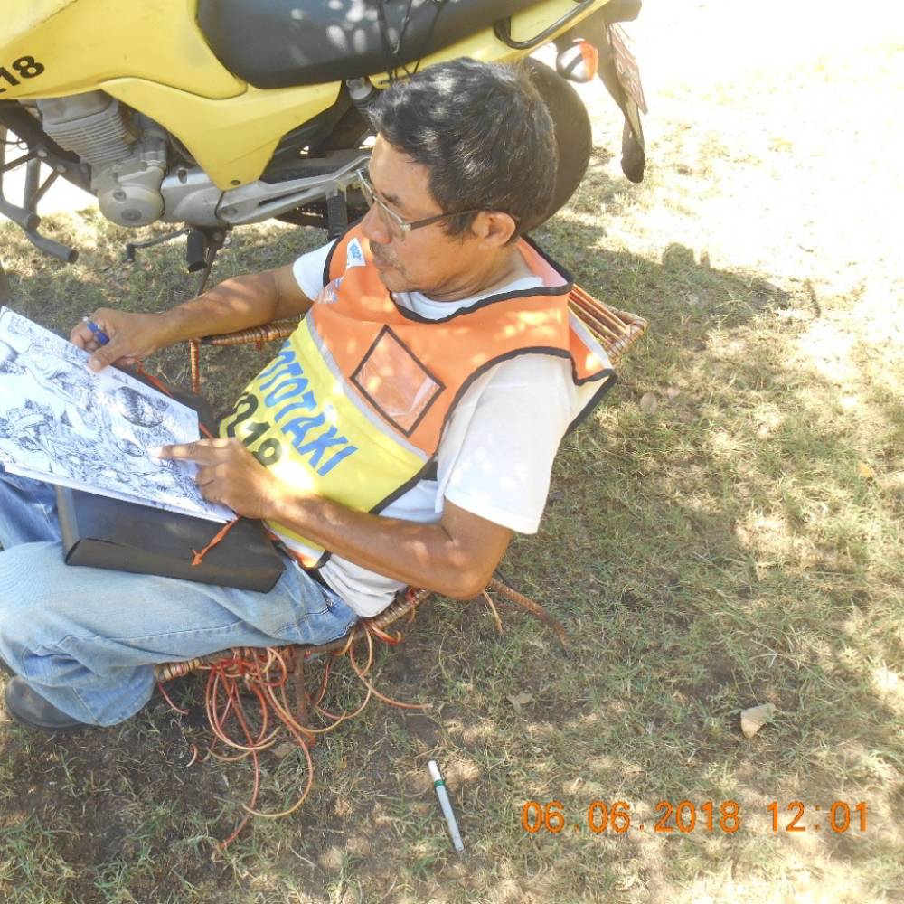 Foto de Luís Celso sentado numa praça. Ele veste o colete laranja de mototaxista e a moto dele está estacionada logo ao lado. Ele está de cabeça baixa desenhando algo que está apoiado nas pernas dele.