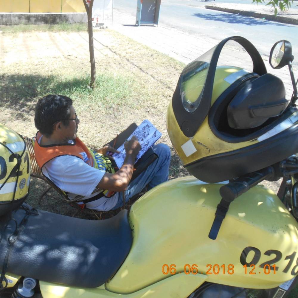 Foto de Luís Celso desenhando na praça enquanto espera alguma corrida de mototaxi. A moto amarela está em destaque.