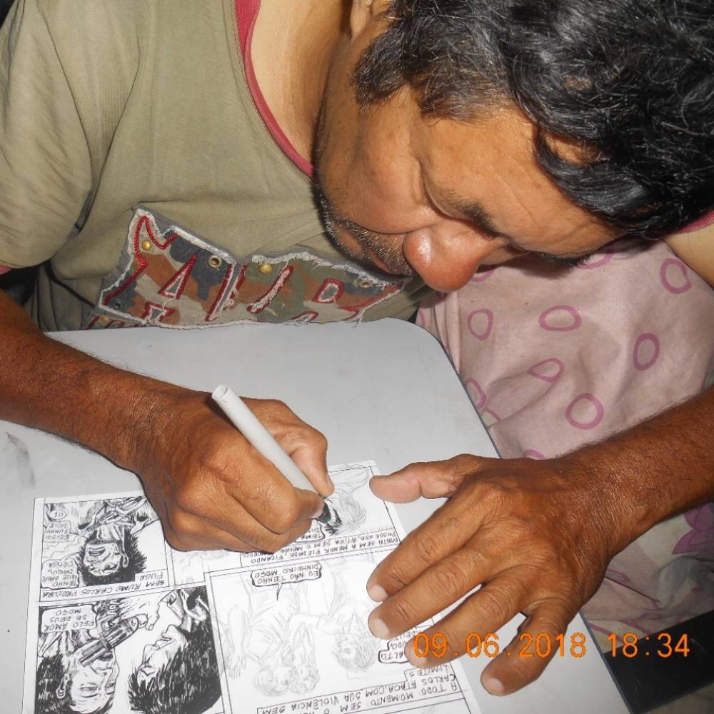 Foto de Luís Celso concentrado desenhando uma página de história em quadrinho. Ele veste uma camisa verde e está de cabeça baixa.