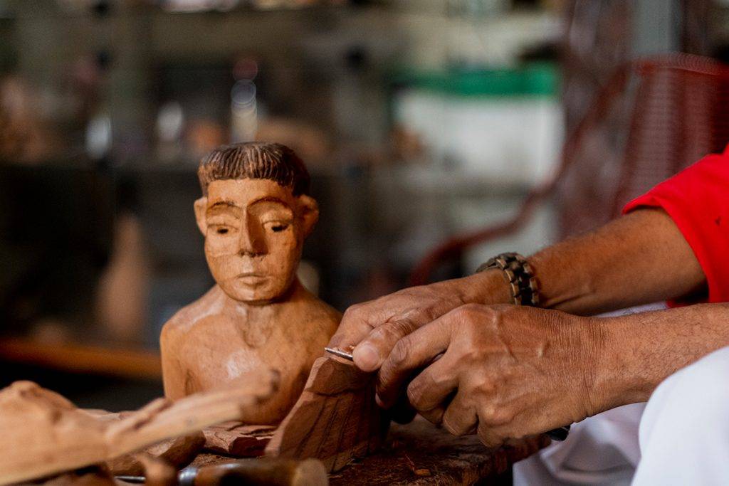 mãos de artesão de idade avançada trabalhando a madeira com uma faca. ao fundo uma imagem de busto feita em madeira.