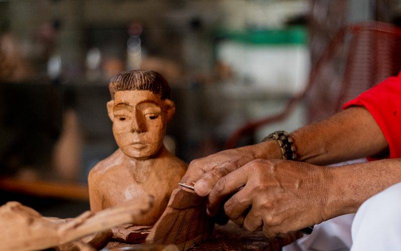 mãos de artesão de idade avançada trabalhando a madeira com uma faca. ao fundo uma imagem de busto feita em madeira.