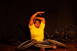 Foto de mulher dançando, ela está sentada no chão, olhos fechados, os braços erguidos, há três rodas de cadeiras de roda sobre as pernas dela.