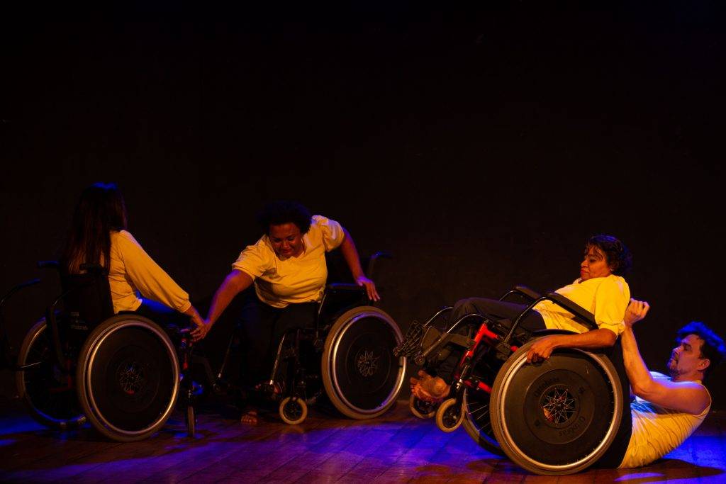 Apresentação da Cia. Dança Eficiente. São quatro pessoas dançando no palco. Elas estão sentadas em cadeiras de rodas, vestem calças pretas e camisas amarelas.