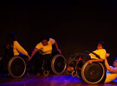 Apresentação da Cia. Dança Eficiente. São quatro pessoas dançando no palco. Elas estão sentadas em cadeiras de rodas, vestem calças pretas e camisas amarelas.