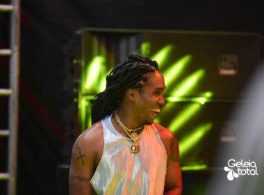Foto do rapper Preto Kedé sorrindo e em cima do palco. “deixa a chuva cair / deixa o céu se abrir / deixa o mundo girar / não só em torno de ti”.