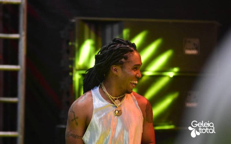 Foto do rapper Preto Kedé sorrindo e em cima do palco. “deixa a chuva cair / deixa o céu se abrir / deixa o mundo girar / não só em torno de ti”.