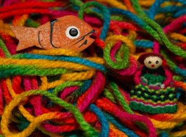 Foto com um pequeno peixe laranja feito de madeira, e uma boneca colorida de tricô, em um fundo feito de vários fios de lã de diversas cores: amarelo, rosa, vermelho, verde, azul. Imagem para representar o Dia Mundial de Conscientização do Autismo.