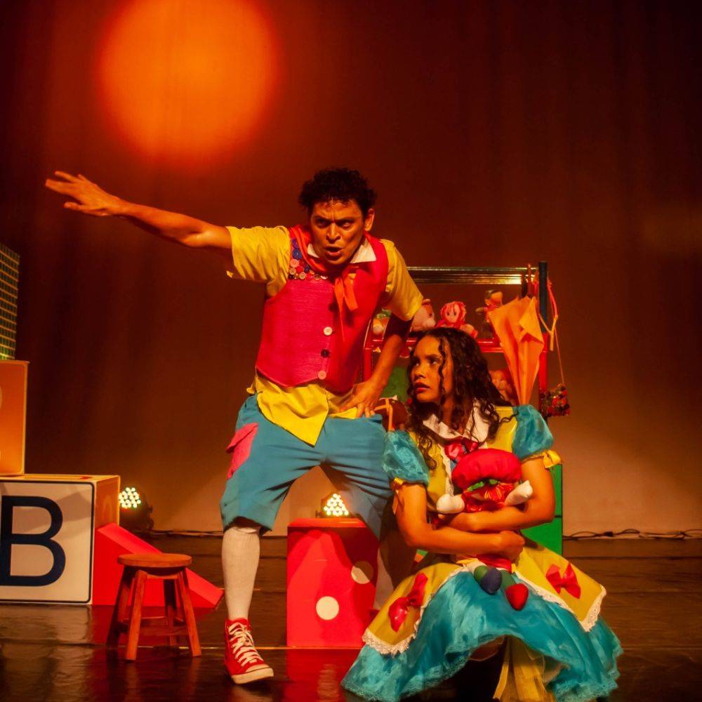 Foto do espetáculo Fábulas. Há dois atores no palco vestindo roupas bem coloridas: vermelho, amarelo, azul. A atriz está sentada no chão abraçada a uma boneca de pano. O ator está em pé ao lado dela e aponta para algo fora de cena.