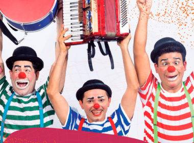 Foto do Grupo Vagão, três palhaços rindo e erguendo instrumentos musicais: um tambor, uma sanfona e um triângulo.