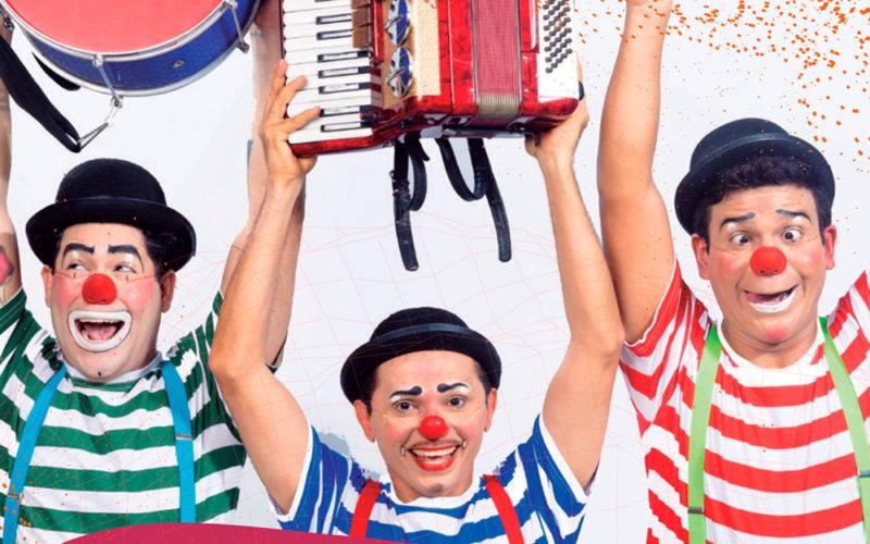 Foto do Grupo Vagão, três palhaços rindo e erguendo instrumentos musicais: um tambor, uma sanfona e um triângulo.