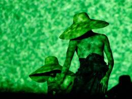 Foto do palco, três dançarinos apresentam o espetáculo Guiança. Sob uma luz verde que simula está debaixo d'água, os três dançarinos chapéus de palha que lhes cobrem os rostos. Foto por Caio Silva.