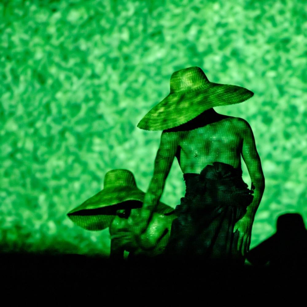 Foto do palco, três dançarinos apresentam o espetáculo Guiança. Sob uma luz verde que simula está debaixo d'água, os três dançarinos chapéus de palha que lhes cobrem os rostos. Foto por Caio Silva.
