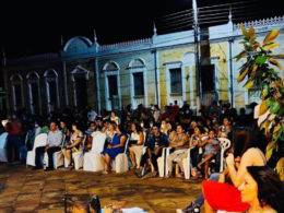 Foto da praça à noite cheia de pessoas sentadas assistindo às apresentações.