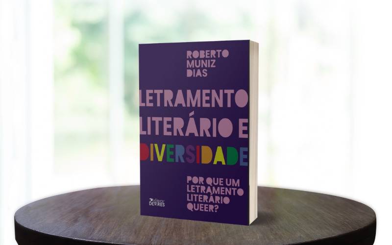 Foto do livro Letramento Literário e Diversidade de autoria de Roberto Muniz Dias. O livro tem a capa roxa e as letras da palavra "diversidade" estão cada uma de uma cor formando um arco-íris. O livro está em cima de uma mesa de madeira escura. Ao fundo uma janela desfocada.