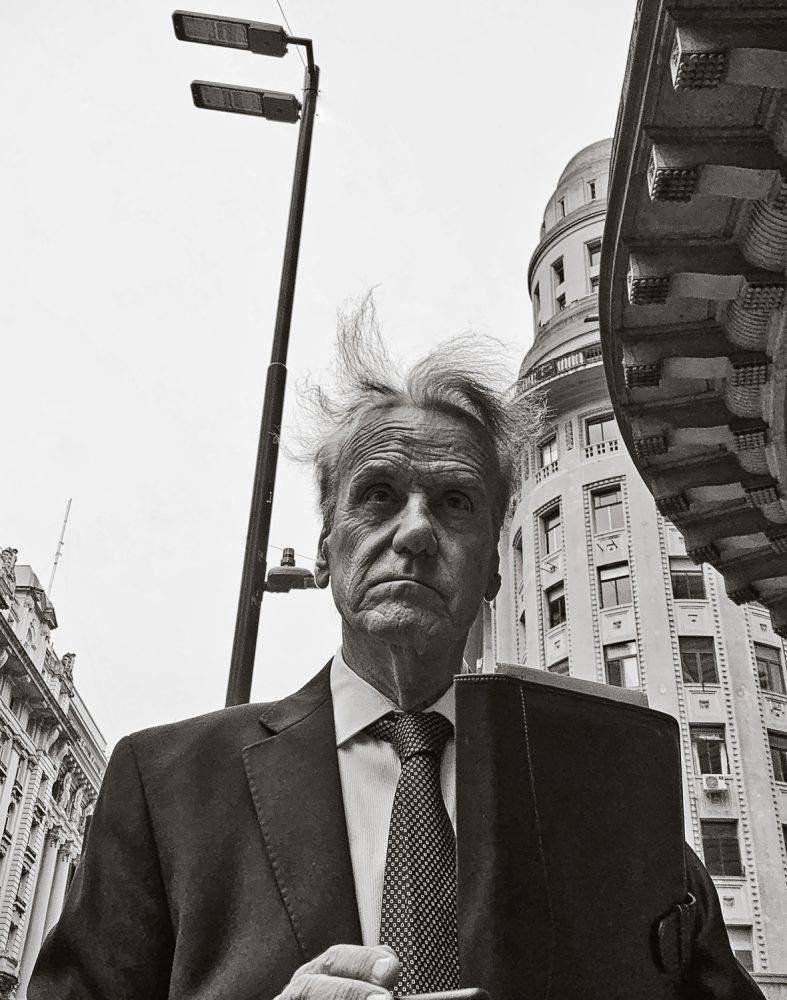 Foto em preto e branco de um senhor de terno com o cabelo bagunçado pelo vento. Ao fundo, a fachada de alguns prédios. Obra do fotógrafo piauiense Adriano Carvalho, premiada no IPPAWARDS.