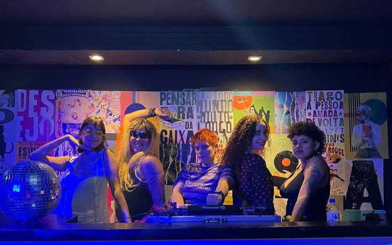 Foto do Boilerage em que aparecem 5 pessoas em uma mesa de DJ, ao fundo vários cartazes cobrem a parede.